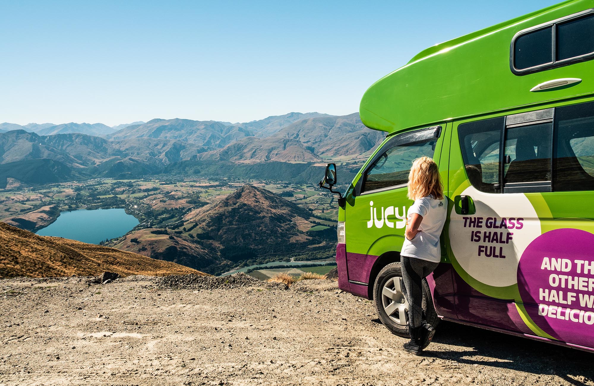 Een meisje leunt tegen een Jucy Condo camper, ze kijkt naar een meer en bergen.
