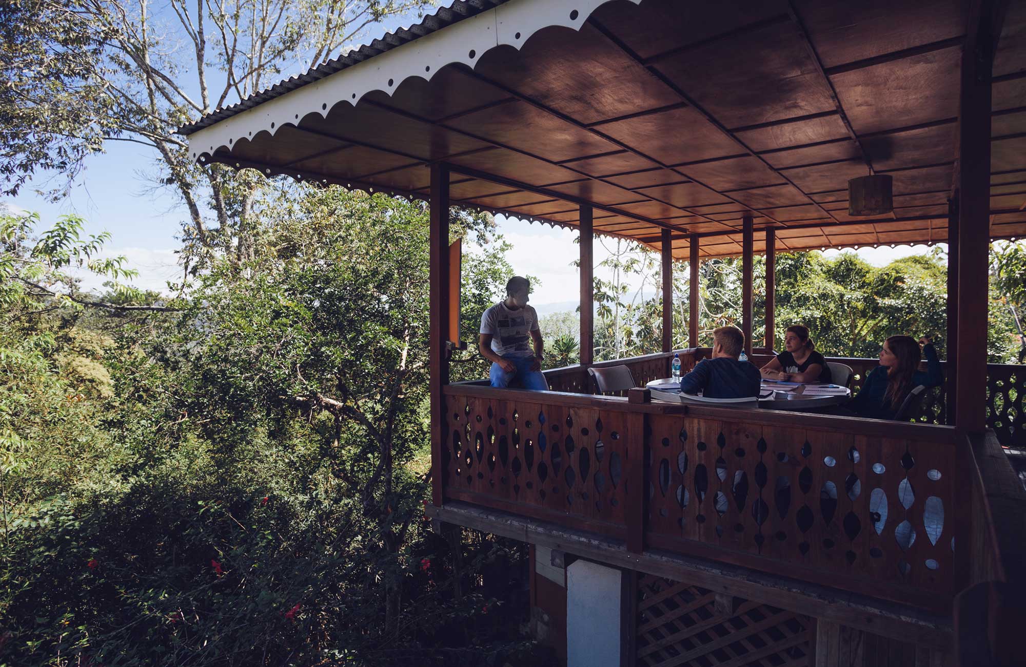 Mensen zitten een rond tafel op een overdekt terras, omgeven door groene natuur