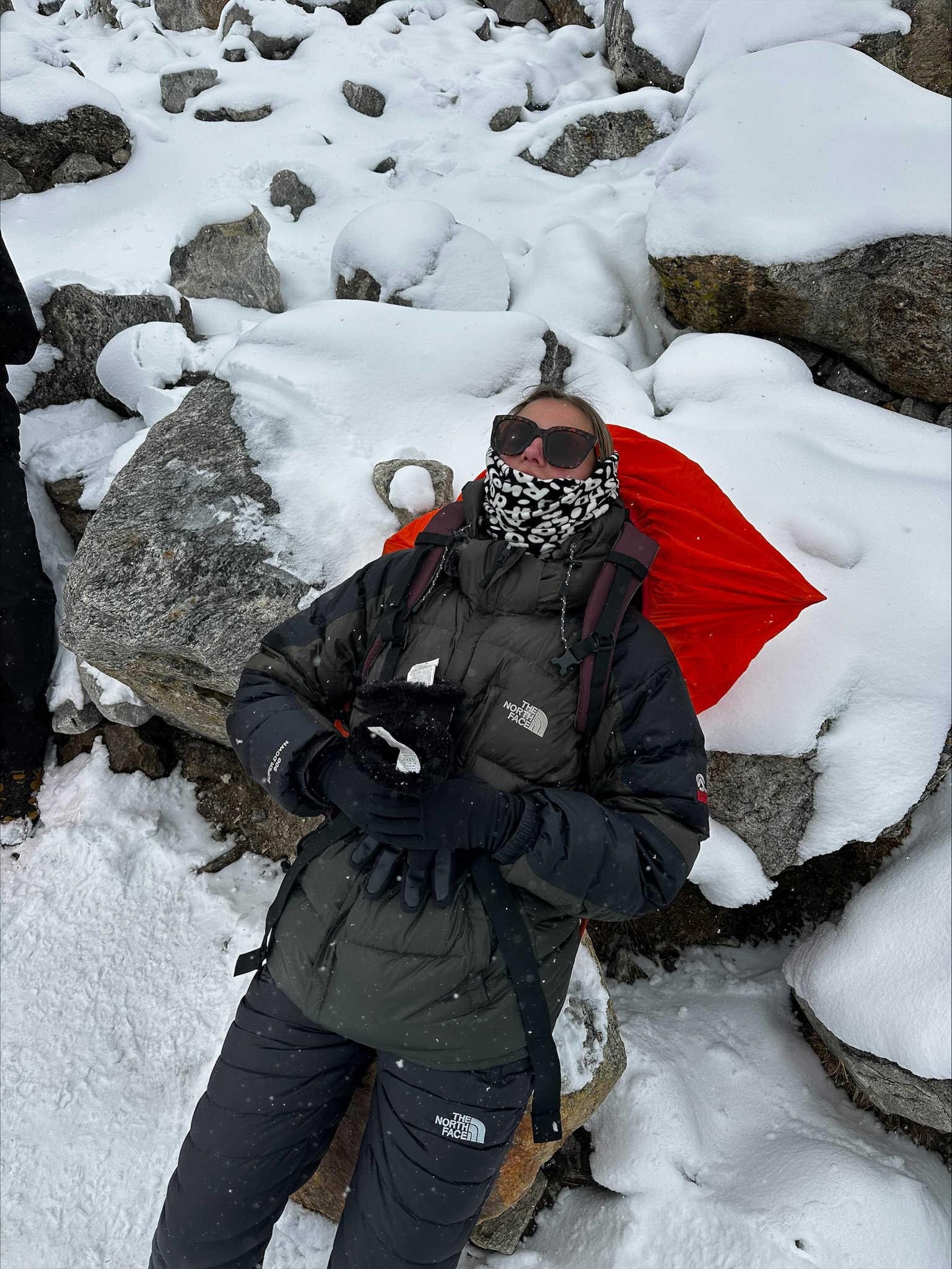 Hiker ligt met zonnebril en warme kleding uit te rusten in de sneeuw met haar rode rugzak als kussen.