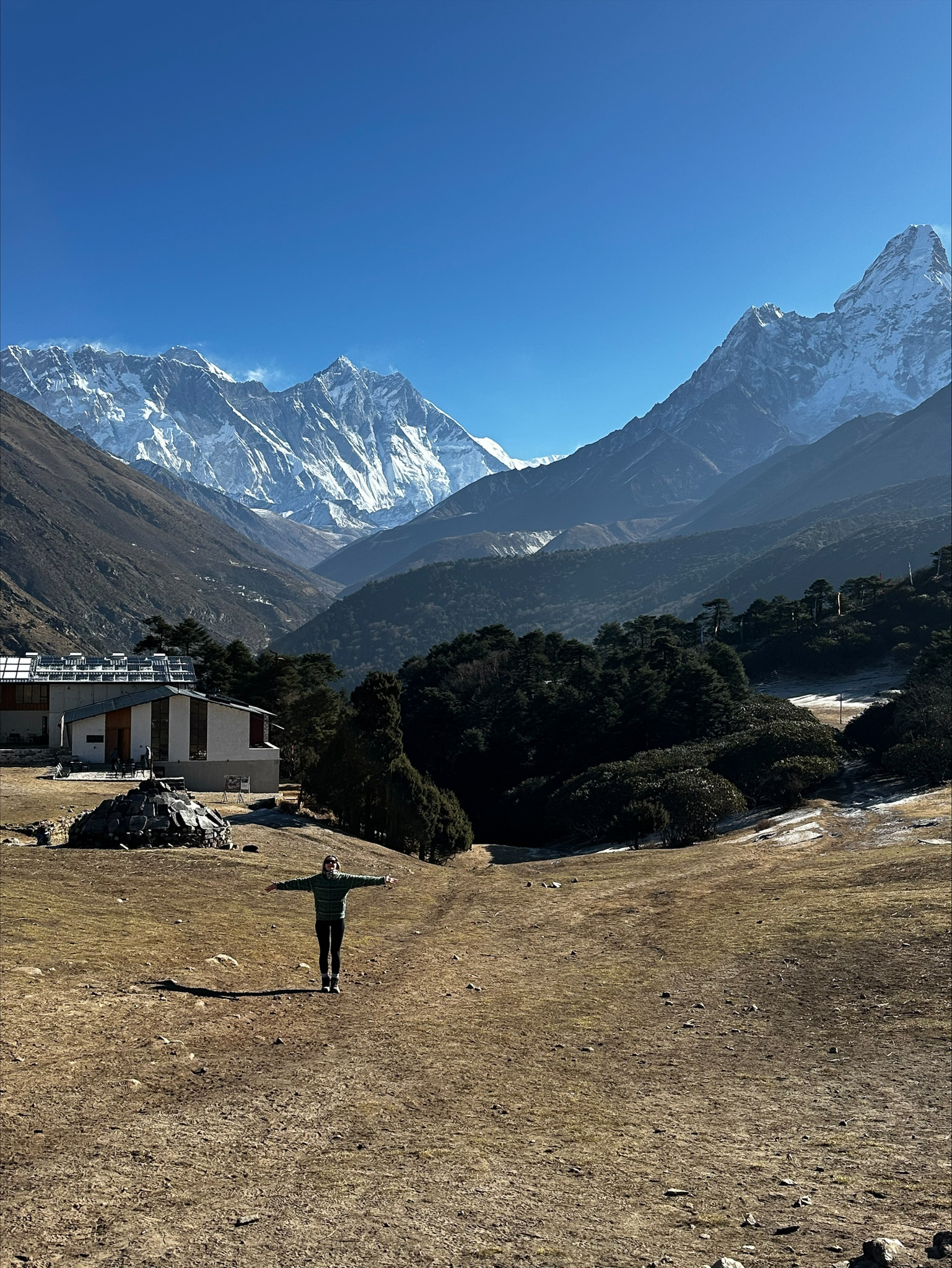 Hiker staat met open armen in een bergdreef met een panoramisch uitzicht op besneeuwde bergtoppen in de Himalaya onder een strakblauwe lucht.